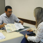 Imagem de um servidor realizando o atendimento de revalidação de diplomas, ele está segurando um diploma , sentado atras de uma mesa enquanto uma moça loira está do outro lado da mesa.