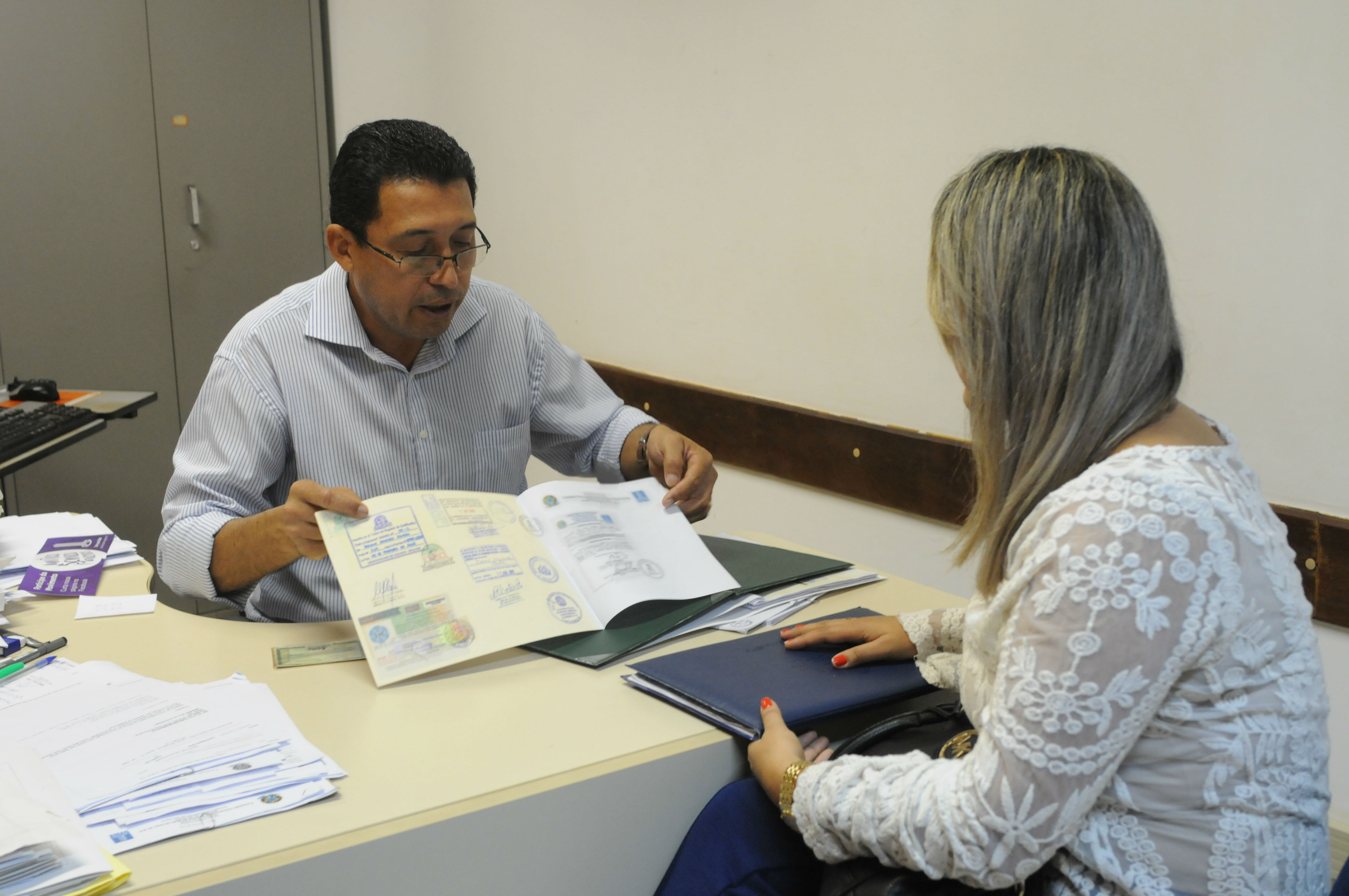 Imagem de um servidor realizando o atendimento de revalidação de diplomas, ele está segurando um diploma , sentado atras de uma mesa enquanto uma moça loira está do outro lado da mesa.