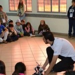 Imagem com um adulto operando um robô. Ele está rodeado de crianças que estão prestando atenção na fala do adulto.