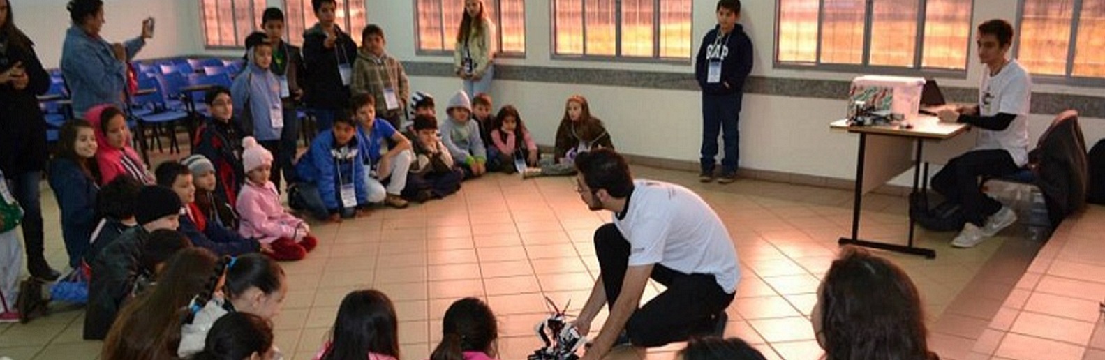 Imagem com um adulto operando um robô. Ele está rodeado de crianças que estão prestando atenção na fala do adulto.