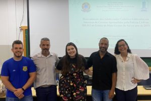 Foto colorida com cinco pessoas em pé na frente de uma mesa e tela de projeção.